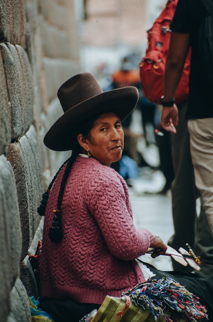 A local woman in traditional attire sits beside a stone wall in Cusco, Peru.